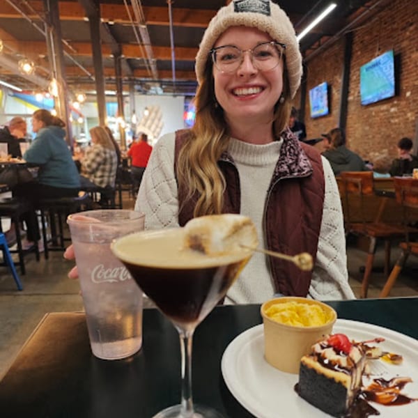 A smiling woman at Oddfellows with a cocktail, cheesecake, and side dish on a table.