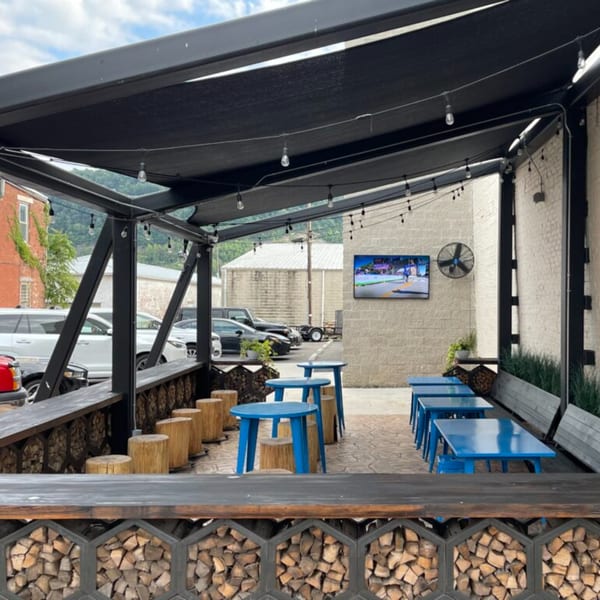Oddfellows' outdoor patio with blue tables, tree stump seats, string lights, and a TV on a brick wall.