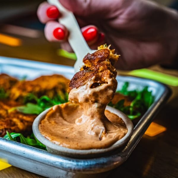 A hand with red nail polish dips a piece of crispy chicken into a creamy sauce.
