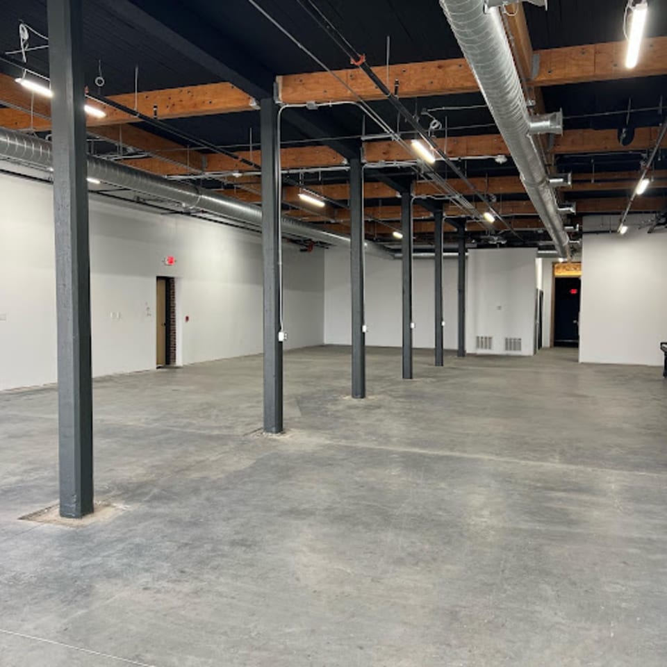 An empty, industrial-style room with concrete floors, white walls, exposed wooden beams, and metal pillars.