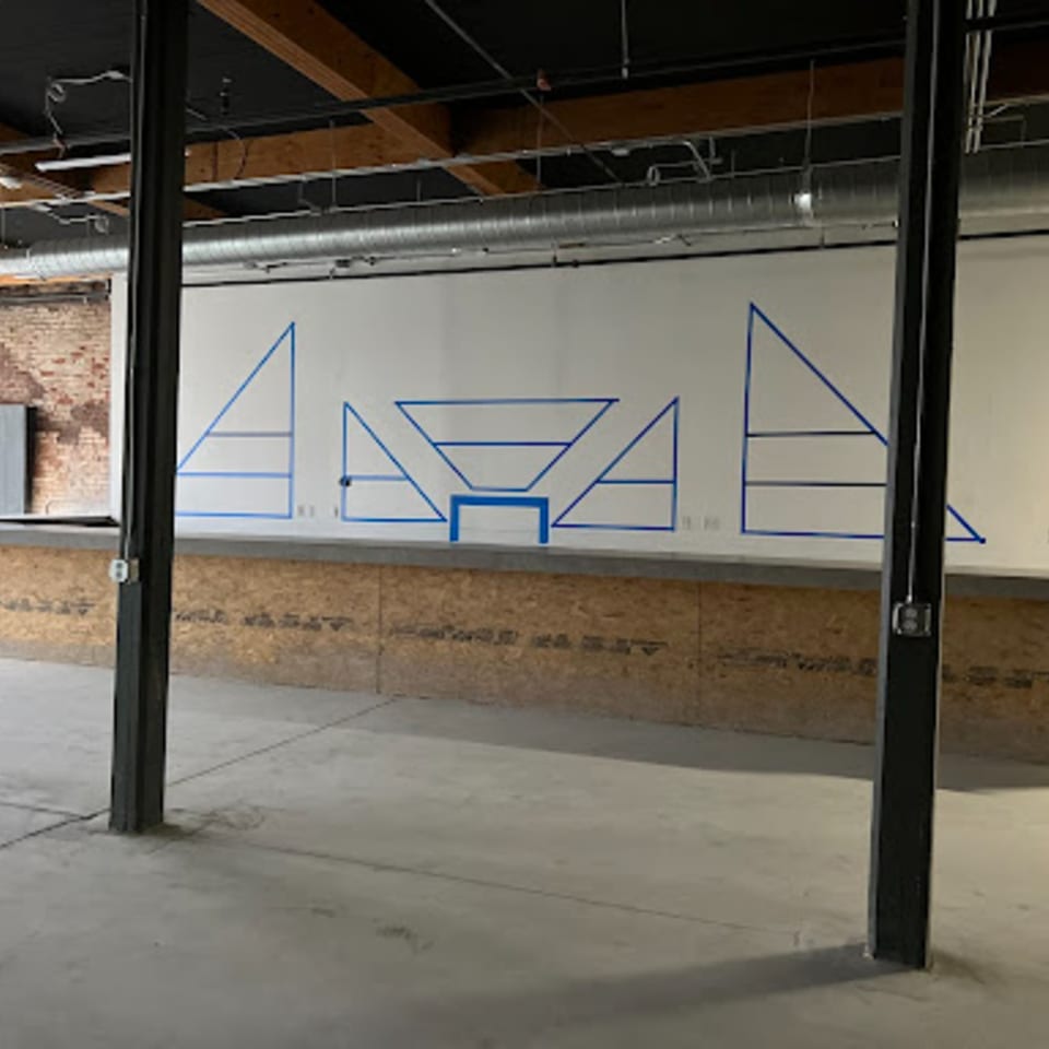 Unfinished restaurant interior with concrete floor, exposed ceiling, and blue tape geometric designs on a white wall.