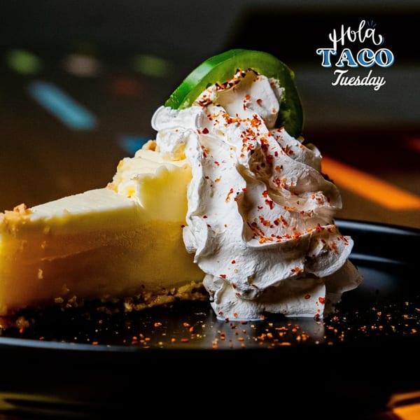 Cheesecake slice topped with whipped cream, chili flakes, and a green pepper slice on a dark plate.