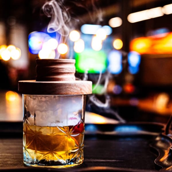 A smoked cocktail in a cut glass with a wooden lid, against a blurred, colorful bar background.