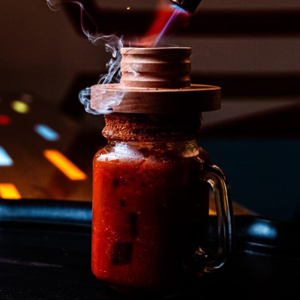 A smoking cocktail in a mason jar with a wooden lid, being torched by a flame.