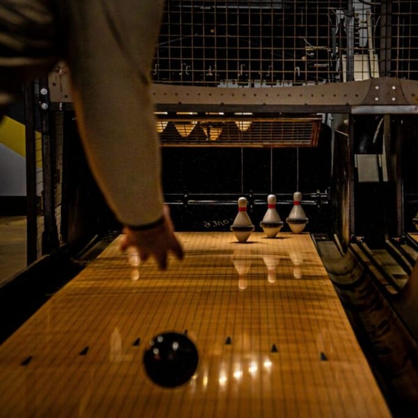 A person bowling, with a black bowling ball rolling towards three white pins on a wooden lane.
