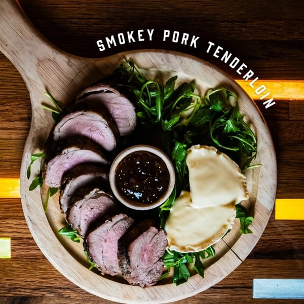Sliced smokey pork tenderloin, arugula, brie cheese, and a dipping sauce on a wooden board.