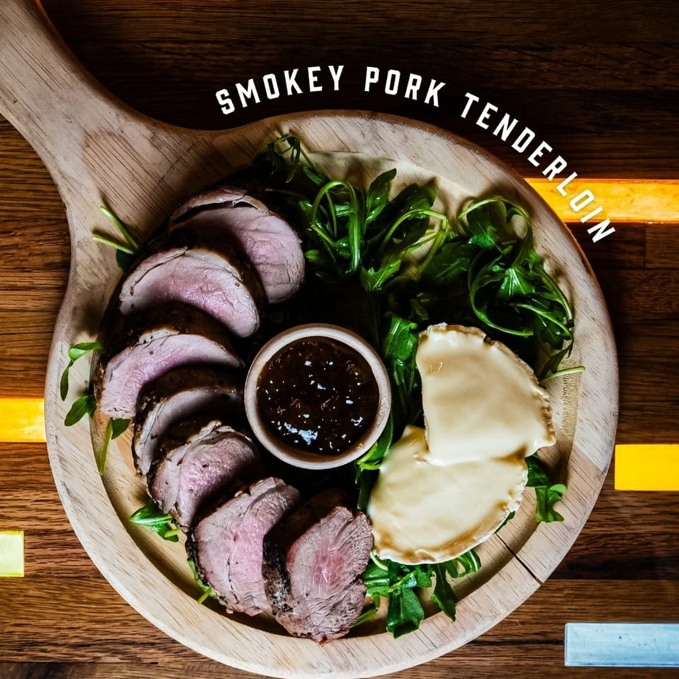 Sliced smokey pork tenderloin, arugula, brie cheese, and a dipping sauce on a wooden board.