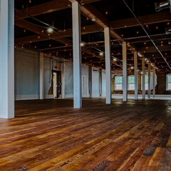 Spacious empty room with wooden floors, exposed beams, string lights, and white support pillars.