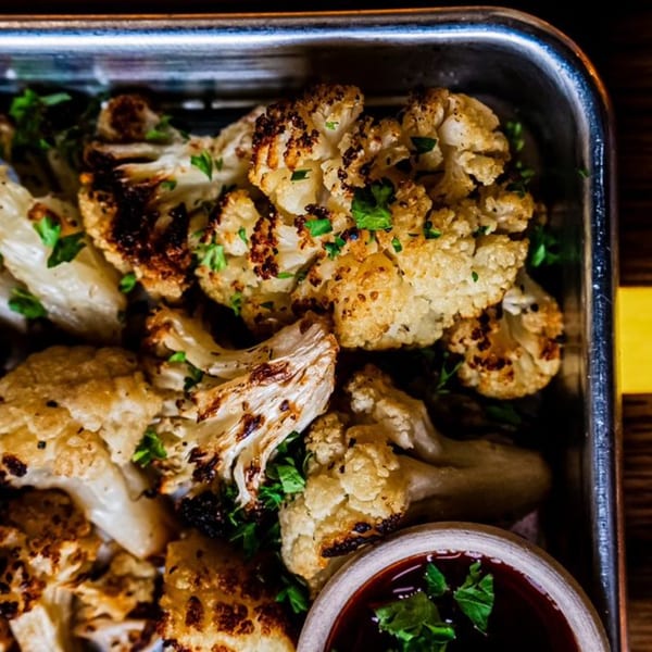 Roasted cauliflower florets with charred edges and fresh parsley in a metal tray with dipping sauce.