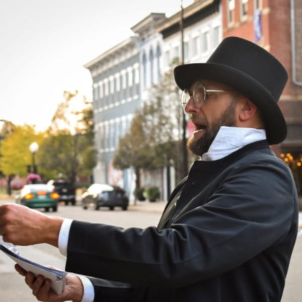 Man in a top hat and glasses, dressed in period clothing, speaking on a street.