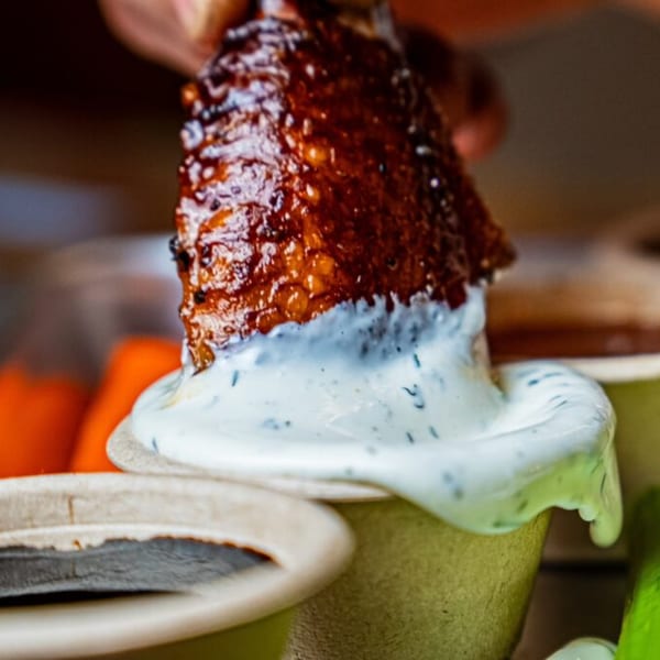 A hand dips a crispy, glazed chicken wing into a cup of creamy ranch dressing.